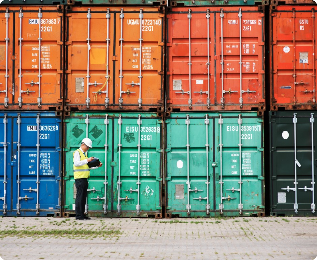 An image of man with a white hard hat and a yellow vest infront of about 8 shipping containers